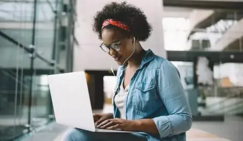 Smiling young African female university student sitting on a campus bench working on a laptop while preparing for an exam